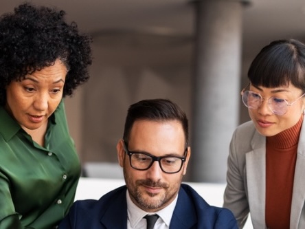 Photo of two women looking over the shoulder of a man and a computer